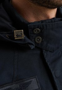 Giacca blu navy con un tessuto strutturato, dotata di un gancio in metallo e bottoni, e di una toppa con un design in rilievo vicino al colletto.
