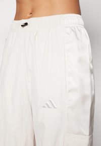 Bílé sportovní kalhoty s elastickým pasem, stahovací šňůrkou a malým reflexním logem na boku, vyrobené z lehkého materiálu.