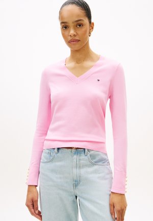 Kobieta ubrana w jasnoróżowy sweter z dekoltem w serek i złotymi guzikami na mankietach oraz jasnoniebieskie, wysokowalne dżinsy, stojąca na białym tle.