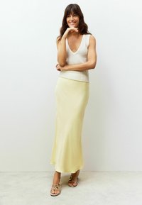 Jupe en satin jaune clair associée à un haut sans manches texturé crème, avec un décolleté en V. Sandales avec des lanières et des accents métalliques.