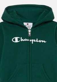 Zelen zip-up kapuca iz mehkih materialov. Vsebuje kenguru žep in velik bel "Champion" logotip na sprednji strani. Nastavljiva kapuca.