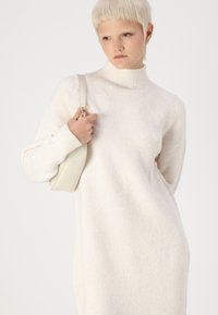 Λευκό πλεκτό φόρεμα με ψηλό λαιμό, μακριά μανίκια και υφασμάτινη επεξεργασία. Το μοντέλο κρατά μια ανοιχτό γκρι δερμάτινη τσάντα με κοντό λουρί.