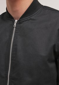 Chaqueta bomber negra con cuello alzado, que presenta un zipper plateado y una textura suave y mate. Diseño sencillo, ajuste clásico.