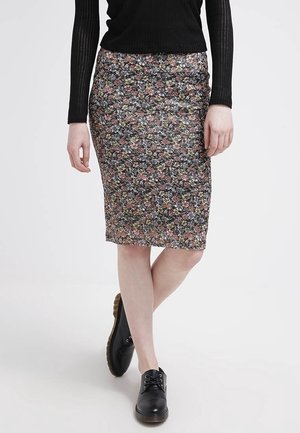 Personne portant une jupe crayon fleurie jusqu'aux genoux, un haut noir à manches longues côtelé et des chaussures noires à lacets, debout avec les jambes croisées.