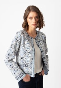 Šviesiai mėlynas raštuotas švarkas su apvaliu kaklu, papuoštas tamsiai mėlynomis detalėmis ir tekstūra, turintis sudėtingus raštus audinyje.