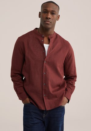 Muškarac obučen u tamnocrvenu košulju s gumbima preko bijele majice i plave traperice, stoji s rukama u džepovima na jednostavnoj pozadini.