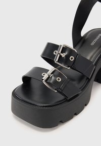 Sandalias de plataforma negras con dos correas anchas y hebillas plateadas, que cuentan con una parte superior de cuero suave y una suela de goma texturizada.