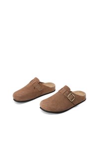 Zoccoli in suede marrone con punta arrotondata, cinturino regolabile e dettaglio in metallo dorato. Presentano una suola in gomma nera per una migliore aderenza.