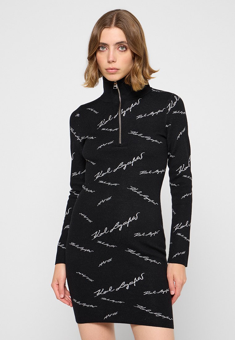 Robe en tricot noire avec un col haut et une fermeture éclair. Présente un motif de texte manuscrit blanc répété sur l'ensemble. Design ajusté.