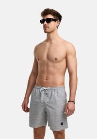 Grå svømmetights med en scalloped mønster, med en hvit snor og en liten svart logo-patch. Båret med svarte solbriller.