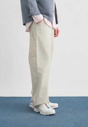 Personne portant un pantalon en velours côtelé beige clair, des baskets blanches avec un logo coloré, une chemise rose clair et une veste grise, debout sur un tapis bleu.