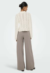 Pull crème en maille ouverte avec manches longues et ourlet à basque, associé à un pantalon large marron clair, présentant une texture lisse et un design élégant.