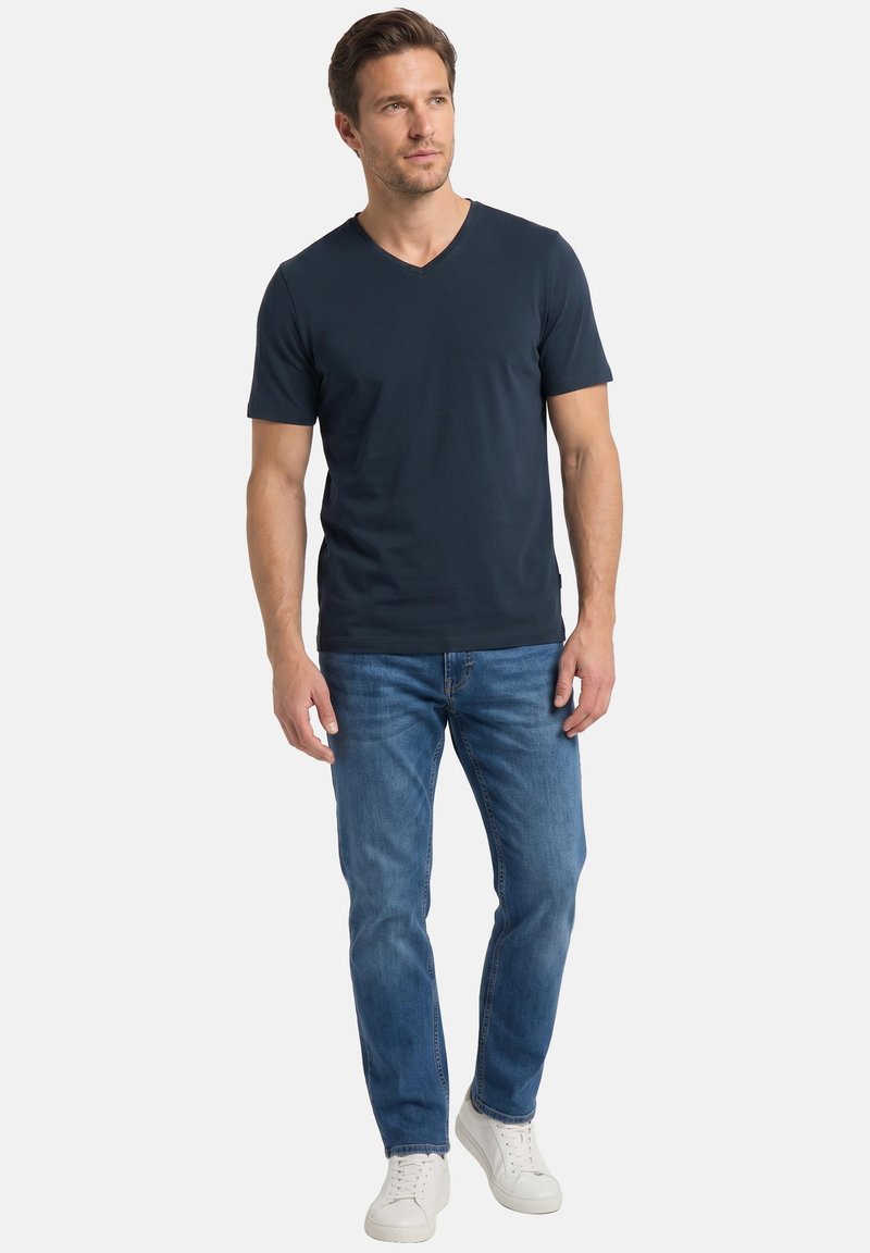 Homme portant un t-shirt bleu marine à col en V, un jean bleu et des baskets blanches, debout les mains détendues sur les côtés.