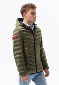 Giacca puffer verde oliva con un design aderente, texture a quadri orizzontali, zip frontale, fodera nera del cappuccio e una toppa rossa con logo sulla manica.