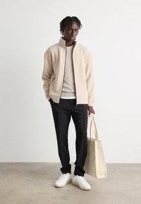 Beige fleece zip-up jacket over a light sweater, paired with black trousers and white sneakers. Carrying a beige tote bag. Simple design.