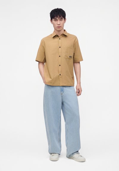 Homme portant une chemise beige à manches courtes boutonnée, un jean bleu clair à jambes larges et des baskets blanches, debout devant un fond uni.