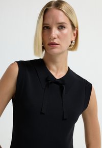 Abito nero senza maniche con colletto rotondo e un fiocco abbinato sullo scollo, realizzato in un tessuto liscio e strutturato con una leggera lucentezza.