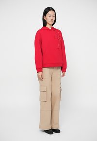 Γυναίκα με μαύρα μαλλιά ντυμένη με κόκκινο φούτερ με ραμμένο φιόγκο και μπεζ φαρδιές παντελόνες cargo, στηριγμένη σε λευκό φόντο.