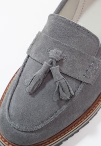Mocassin en suede gris avec des détails en pompons, bout arrondi, broderies décoratives et une semelle extérieure brune contrastante avec un motif de relief visible.