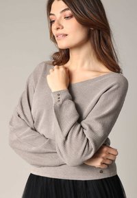 Pull en beige à côtes avec un large col, des épaules tombantes et des accents de boutons sur les poignets. Texture douce, associé à une jupe en tulle noire.