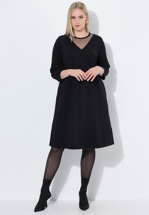 Schwarzes Kleid mit V-Ausschnitt und transparenter Netz-Einlage. Lange Ärmel, knielang. Glattes Material, dezenter A-Linien-Schnitt. Kombiniert mit transparenten Strumpfhosen und knöchelhohen Stiefeln.