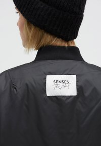 Black bomber jacket featuring a smooth, glossy fabric and a ribbed collar. Displays a white label with "Senses The Label" branding on the back.