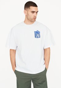 Camiseta blanca de algodón de gran tamaño con un estampado gráfico azul en el bolsillo. Manga corta, cuello redondo, corte relajado. Combinada con pantalones verdes.