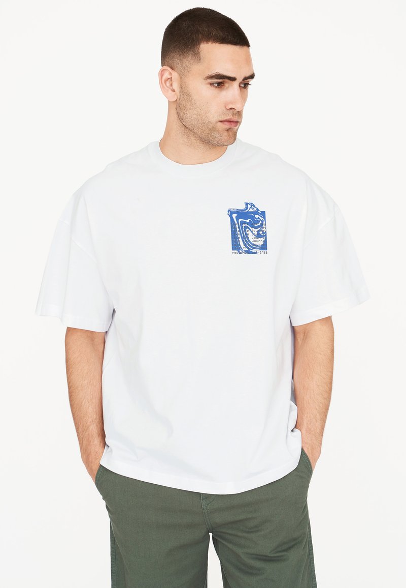 Camiseta blanca de algodón de gran tamaño con un estampado gráfico azul en el bolsillo. Manga corta, cuello redondo, corte relajado. Combinada con pantalones verdes.