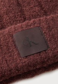Bordeaux teksturert strikket lue med et rektangulært brunt merke preget med bokstavene "cK." Mykt, varmt materiale med en lodden overflate.