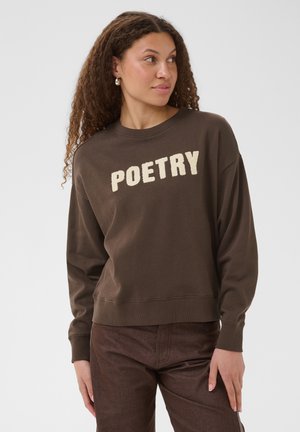 Kvinde med krøllet hår iført en brun sweatshirt med ordet "POETRY" i fede hvide bogstaver, ser til siden foran en ensfarvet baggrund.
