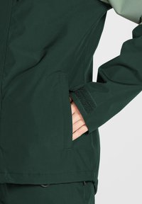 Dunkelgrüne Jacke mit einem Reißverschluss vorne, einem Klettverschlussfach und strapazierfähigem Material. Geschmeidige Textur mit einer figurbetonten Passform und geformten Bündchen.