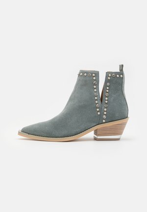 Bottine en suede grise avec un bout pointu, ornée de clous argentés le long des coutures latérales, et un talon en bois empilé avec un accent métallique.