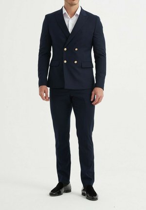 Costume navy à double boutonnage avec des boutons dorés, pantalon assorti et chemise blanche. Le tissu semble lisse ; le design présente une coupe élégante.
