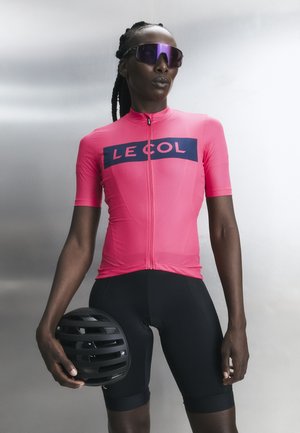 Ποδηλάτης που φοράει ροζ φανέλα LE COL και μαύρο σορτς, κρατώντας μαύρο κράνος, φορά ανακλαστικά μωβ γυαλιά ηλίου, στέκεται μπροστά σε απλό φόντο.