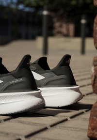 Scarpe sportive nere e grigie con una parte superiore testurizzata, una soletta bianca e il marchio 'ULTRABOOST' sul tallone. Design imbottito con una forma aerodinamica.