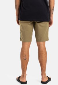 Shorts en coton beige avec une coupe droite, dotés d'une ceinture plate et de poches latérales, portés avec des tongs noires, montrant un tatouage sur la jambe.