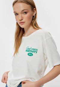 Hvid bomulds t-shirt med grøn trykt tekst "Good Vibes Positivity '90s." Løs pasform med korte ærmer og rund hals.