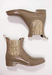 Botas de tobillo de goma marrones con un acabado brillante, paneles laterales de purpurina texturizada y tiradores para facilitar su uso. Diseño de tacón plano con punteras redondeadas.