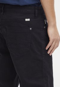 Pantalones de algodón negros que presentan un diseño de cinco bolsillos, con una etiqueta de logo visible en la parte posterior y una silueta recta. Cintura elástica.