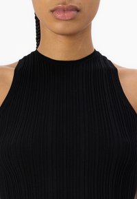 Primer plano de una persona que lleva una camiseta negra de canalé sin mangas con un escote alto, mostrando los hombros y los labios contra un fondo blanco.