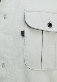 Lichtgrijze shirtstof met een klepzak, knoopdetails en een klein zwart etiket met de letter "J." Zijopeningen nabij de halslijn.