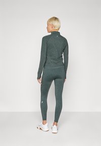 Ensemble de sport vert foncé comprenant un haut à manches longues avec un col haut et un legging moulant. Les deux pièces ont une texture scintillante subtile.