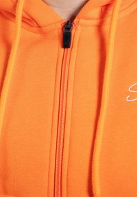 Haut à zip orange en tissu doux. Présente une fermeture éclair noire et un petit détail brodé sur le côté droit. Texturé avec des accents côtelés.