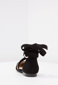 Sandalo flat nero con cinturini in suede, fiocco annodato alla caviglia e fibbia argentata, mostrato da dietro su una superficie bianca.
