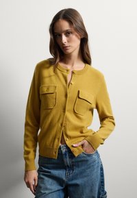 Cardigan giallo con scollo rotondo, due tasche frontali e chiusura con bottoni, realizzato in un tessuto morbido. Abbinato a un paio di jeans a vita alta.