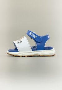 Παιδικό ανοιχτό πέδιλο σε μπλε και άσπρο με λουράκια Velcro, λευκή σόλα και λογότυπο "MOSCHINO" στο λουράκι του αστραγάλου.