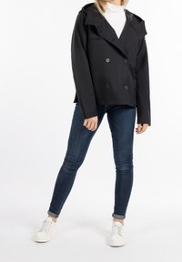 Chaqueta negra impermeable con un diseño de doble botonadura, gran cuello y capucha. Presenta una tela suave y detalles de botones. Combinada con jeans de mezclilla y zapatillas blancas.