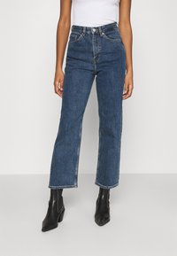 Person som bär blåa jeans med hög midja och raka ben samt svarta ankelboots med klack, med en vit topp mot en enfärgad bakgrund.