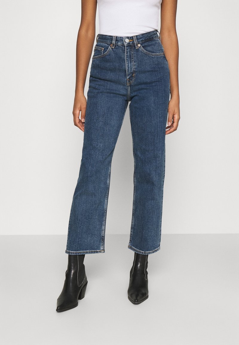 Person som bär blåa jeans med hög midja och raka ben samt svarta ankelboots med klack, med en vit topp mot en enfärgad bakgrund.