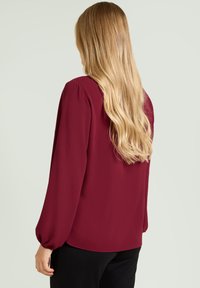 Bordowa bluzka z długim rękawem o gładkiej fakturze i bufiaste ramiona, z luźnym krojem i delikatnym drapowaniem z tyłu.